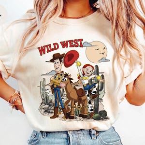 May include: Ivory-colored t-shirt featuring a Wild West design. The graphic includes cartoon characters, a horse, cacti, and a sun. The text "WILD WEST" is printed in red. The shirt is a casual wear item.