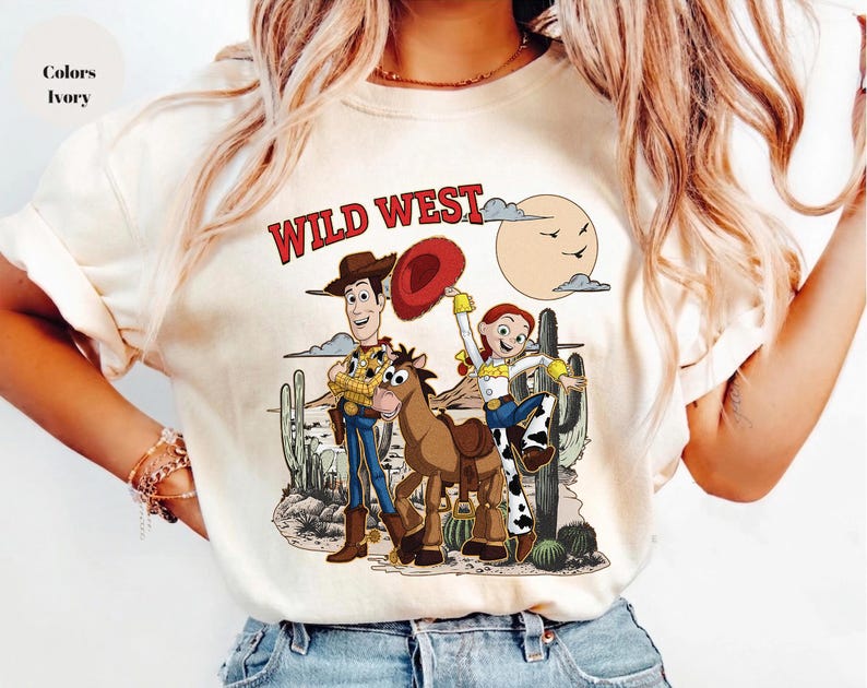 May include: Ivory-colored t-shirt featuring a Wild West design. The graphic includes cartoon characters, a horse, cacti, and a sun. The text "WILD WEST" is printed in red. The shirt is a casual wear item.