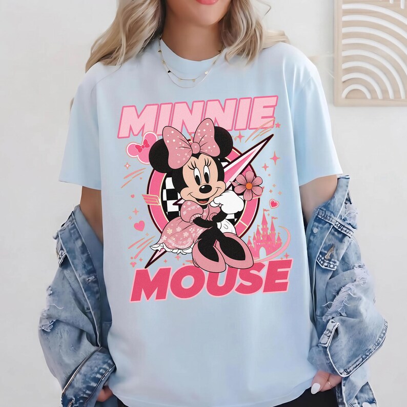 May include: Light blue t-shirt featuring a Minnie Mouse graphic. The graphic includes the text "MINNIE MOUSE" in pink, along with an illustration of Minnie Mouse in a pink dress, holding a flower. The design incorporates hearts, stars, and a castle.