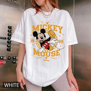 May include: White t-shirt featuring a graphic of Mickey Mouse in a dynamic pose. The design includes the words "Mickey Mouse" in yellow, along with the year "1928". The Disney logo is also visible. The word "WHITE" is printed at the bottom.