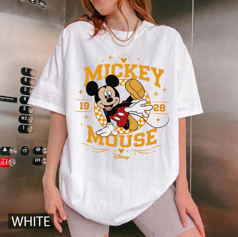 May include: White t-shirt featuring a graphic of Mickey Mouse in a dynamic pose. The design includes the words "Mickey Mouse" in yellow, along with the year "1928". The Disney logo is also visible. The word "WHITE" is printed at the bottom.