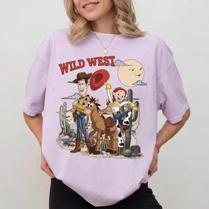 May include: A light purple t-shirt featuring a cartoon illustration of Woody, Jessie, and a horse in a Wild West scene. The text "WILD WEST" is printed in red above the characters. The design includes cacti, a sun, and clouds.