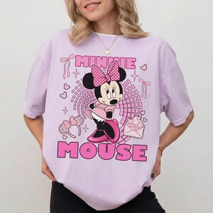 May include: A lilac t-shirt with a Minnie Mouse design. The design shows Minnie Mouse in a pink dress and bow, with the words "MINNIE MOUSE" in pink. Pink hearts, bows, and stars are also included in the design.