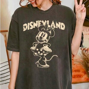May include: Dark gray t-shirt featuring the word "DISNEYLAND" in a distressed font above a Minnie Mouse graphic. The Minnie Mouse illustration is in a vintage style, with a polka-dot skirt and bow.