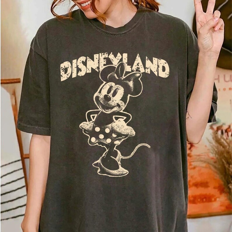 May include: Dark gray t-shirt featuring the word "DISNEYLAND" in a distressed font above a Minnie Mouse graphic. The Minnie Mouse illustration is in a vintage style, with a polka-dot skirt and bow.