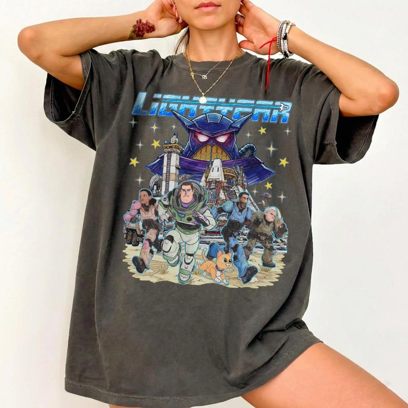 May include: Dark gray t-shirt featuring a colorful graphic of characters from the movie "Lightyear". The design includes Buzz Lightyear, Zurg, and other characters running across a sandy landscape with a starry background. The word "Lightyear" is printed across the top.