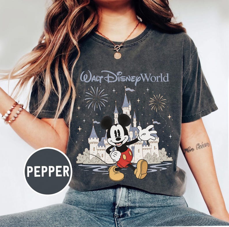 May include: Dark gray t-shirt featuring Mickey Mouse in front of Cinderella's castle with fireworks and the text "Walt Disney World". Mickey is wearing red shorts and yellow shoes. The shirt has a vintage, distressed look.