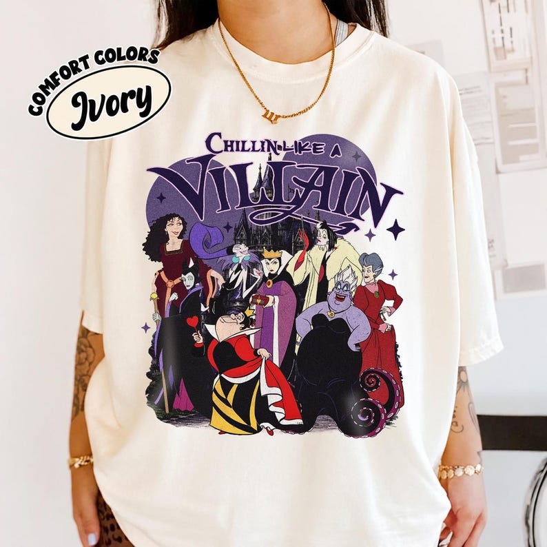 K&ouml;nnte beinhalten: Ein elfenbeinfarbenes T-Shirt mit einem Grafikdruck verschiedener animierter B&ouml;sewichte und dem Text "CHILLIN LIKE A VILLAIN" in lila Schrift dar&uuml;ber. Das Design zeigt Figuren in bunten Outfits vor einer dunklen Schlosskulisse.