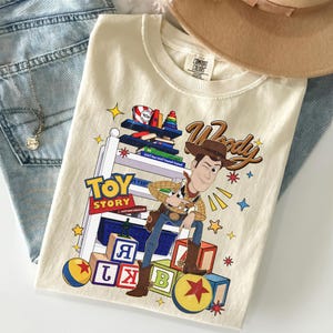 K&ouml;nnte beinhalten: Cremefarbenes T-Shirt mit der Figur Woody aus Toy Story. Die Grafik enth&auml;lt die W&ouml;rter "Toy Story" und "Woody" sowie bunte Bl&ouml;cke, Sterne und ein Spielzeugregal. Das T-Shirt liegt gefaltet auf einer Jeans.