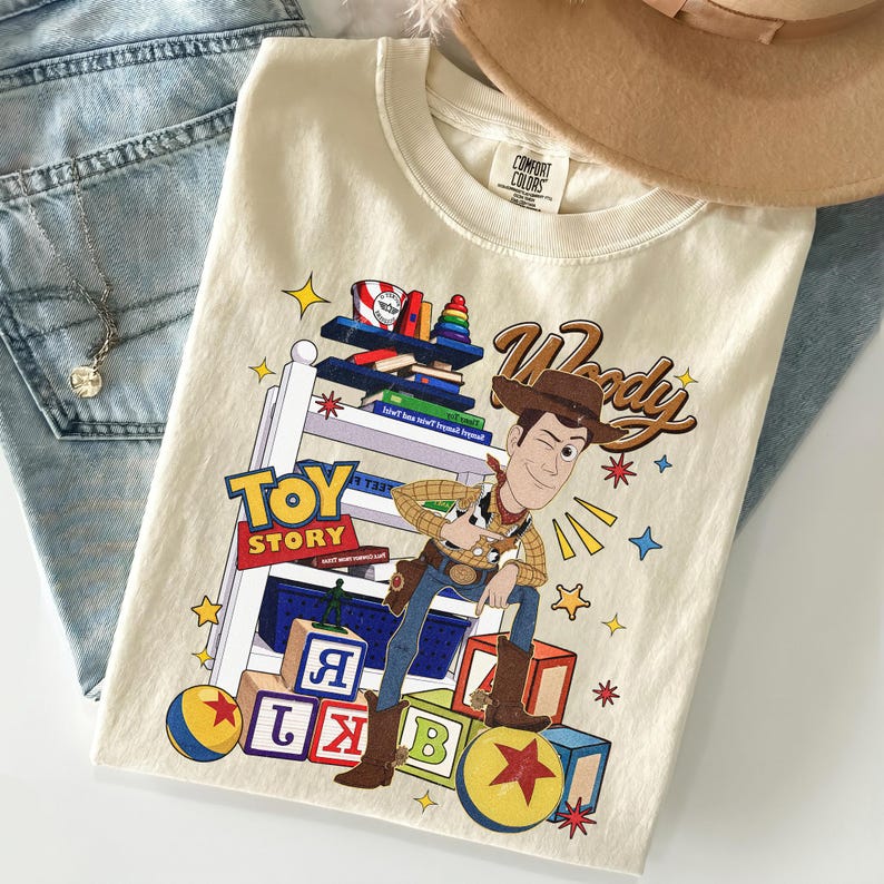 K&ouml;nnte beinhalten: Cremefarbenes T-Shirt mit der Figur Woody aus Toy Story. Die Grafik enth&auml;lt die W&ouml;rter "Toy Story" und "Woody" sowie bunte Bl&ouml;cke, Sterne und ein Spielzeugregal. Das T-Shirt liegt gefaltet auf einer Jeans.