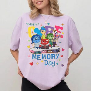 May include: A lilac t-shirt with characters from the animated film "Inside Out." The shirt displays the text "Today is a CORE MEMORY Day" with colourful graphics of the characters and decorative elements.
