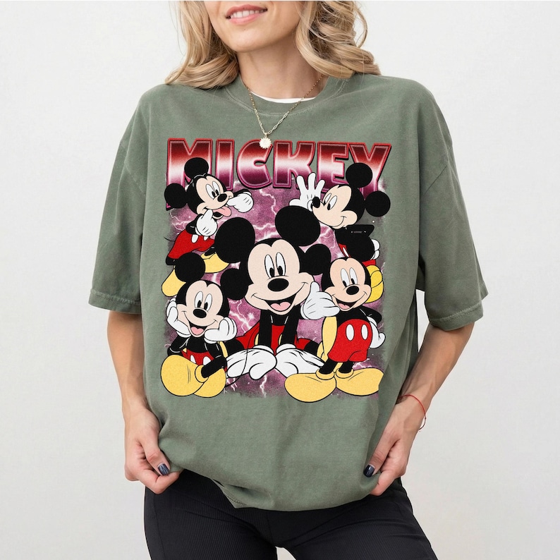 Puede incluir: Camiseta verde salvia con un gr&aacute;fico de Mickey Mouse en varias poses. La palabra "MICKEY" se muestra en una fuente roja y blanca encima del personaje. El dise&ntilde;o incluye un fondo de rayo p&uacute;rpura.