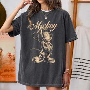 Puede incluir: Una camiseta de manga corta gris carb&oacute;n con un estampado gr&aacute;fico en beige de Mickey Mouse vistiendo una sudadera con capucha y zapatillas. La palabra Mickey est&aacute; impresa en una fuente cursiva sobre la ilustraci&oacute;n en la parte frontal.