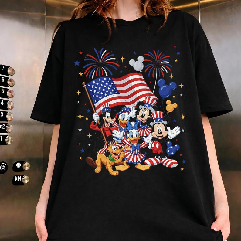 May include: Black t-shirt featuring a patriotic design with Disney characters. The graphic includes Mickey Mouse, Minnie Mouse, Donald Duck, and Pluto, along with an American flag, fireworks, and stars.