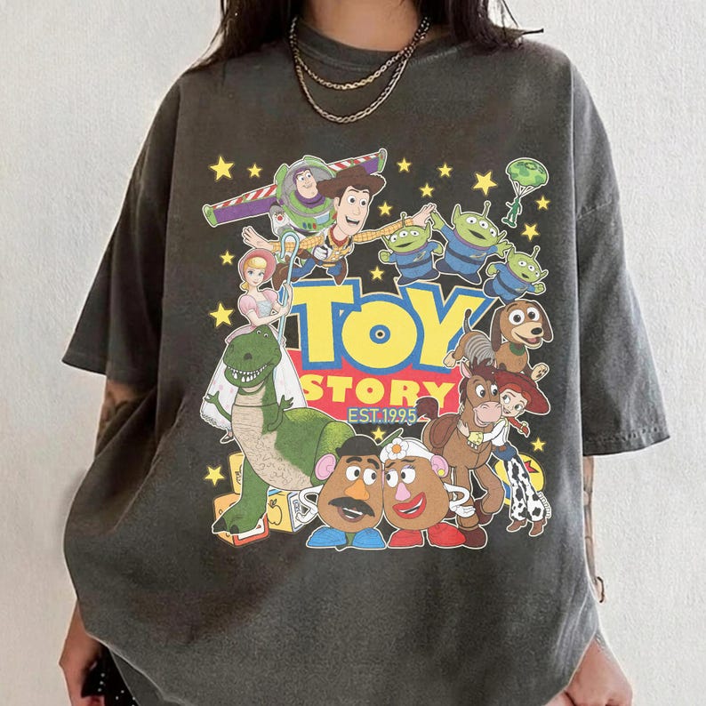 Pu&ograve; includere: T-shirt grigio scuro con una grafica colorata dei personaggi del film "Toy Story". Il design include Woody, Buzz Lightyear e altri personaggi, con le parole "Toy Story" e "Est. 1995" in un font giocoso. La maglietta ha un effetto vintage.