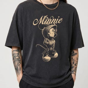 May include: A charcoal black short-sleeve t-shirt featuring a beige graphic print of Minnie Mouse wearing a hooded sweatshirt and sneakers. The word Minnie is printed in a cursive script above the character. The shirt has a crew neckline.