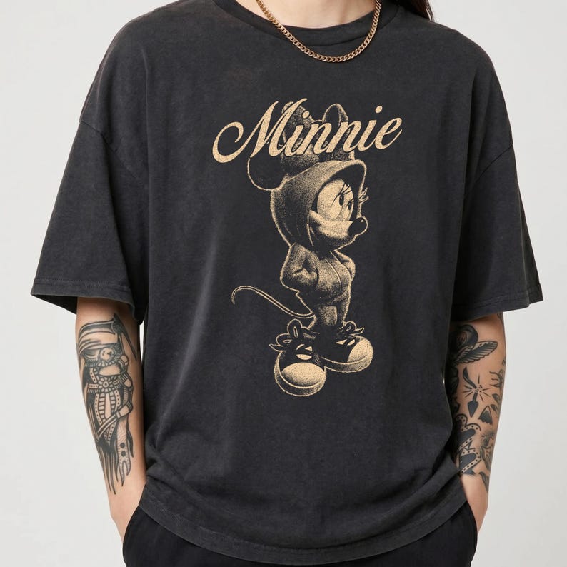 May include: A charcoal black short-sleeve t-shirt featuring a beige graphic print of Minnie Mouse wearing a hooded sweatshirt and sneakers. The word Minnie is printed in a cursive script above the character. The shirt has a crew neckline.
