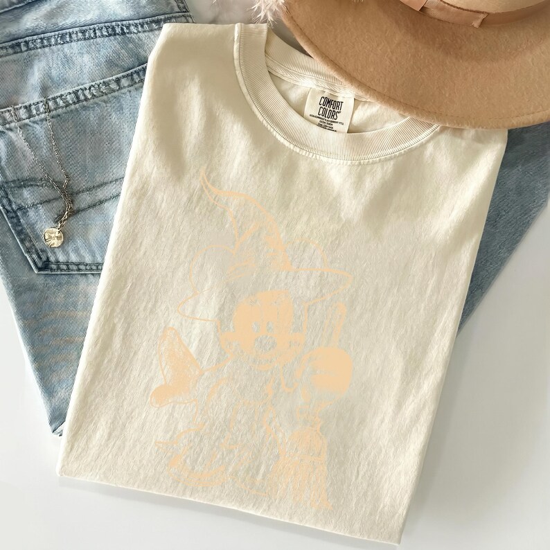 Puede incluir: Camiseta color crema con un dise&ntilde;o gr&aacute;fico color melocot&oacute;n. La camiseta est&aacute; doblada y colocada encima de unos vaqueros azules. Un sombrero color canela es parcialmente visible en la esquina superior derecha. La camiseta tiene una etiqueta Comfort Colors.