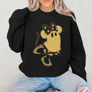 Pode incluir: Camisola preta com um design dourado brilhante de um rato de desenho animado com la&ccedil;o, vestido e sapatos. O rato tem uma cauda longa e est&aacute; em pose de dan&ccedil;a. A camisola &eacute; de estilo casual.