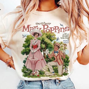 May include: Ivory-colored t-shirt featuring a vintage illustration of Mary Poppins. The graphic includes the film's title, characters, and the text "with a magic tote bag PAPER DOLLS". The design showcases a classic, whimsical aesthetic.