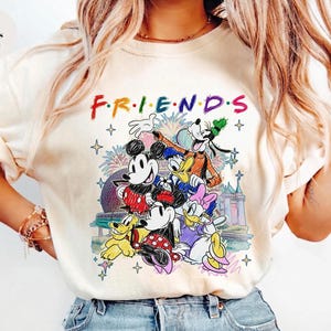 May include: Ivory-colored t-shirt featuring the word "FRIENDS" in colorful letters above a graphic of Disney characters, including Mickey Mouse, Minnie Mouse, Donald Duck, and Goofy. The graphic has a castle and fireworks in the background.