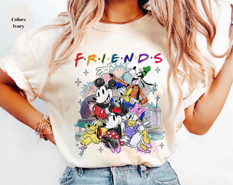 May include: Ivory-colored t-shirt featuring the word "FRIENDS" in colorful letters above a graphic of Disney characters, including Mickey Mouse, Minnie Mouse, Donald Duck, and Goofy. The graphic has a castle and fireworks in the background.