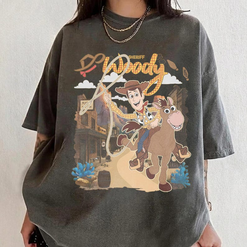 May include: Dark gray t-shirt featuring a graphic of Sheriff Woody from Toy Story riding a horse, with the word "Woody" and "Sheriff" in a western-style font. The design includes a rope and a cityscape background.