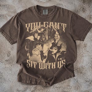 May include: Brown t-shirt featuring a graphic of Disney villains with the text "YOU CAN'T SIT WITH US". The design includes characters like Maleficent and the Evil Queen. The shirt has a vintage, distressed look.