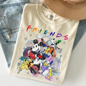 May include: Cream-colored t-shirt featuring the word "FRIENDS" in rainbow colors and Disney characters. The graphic includes Mickey Mouse, Minnie Mouse, Donald Duck, Daisy Duck, Goofy, and Pluto. The shirt is folded on top of blue jeans.