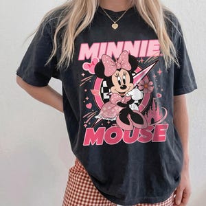 May include: Black t-shirt featuring a graphic of Minnie Mouse in a pink dress and bow. The words "MINNIE MOUSE" are printed in pink. The design includes hearts, stars, and a castle.