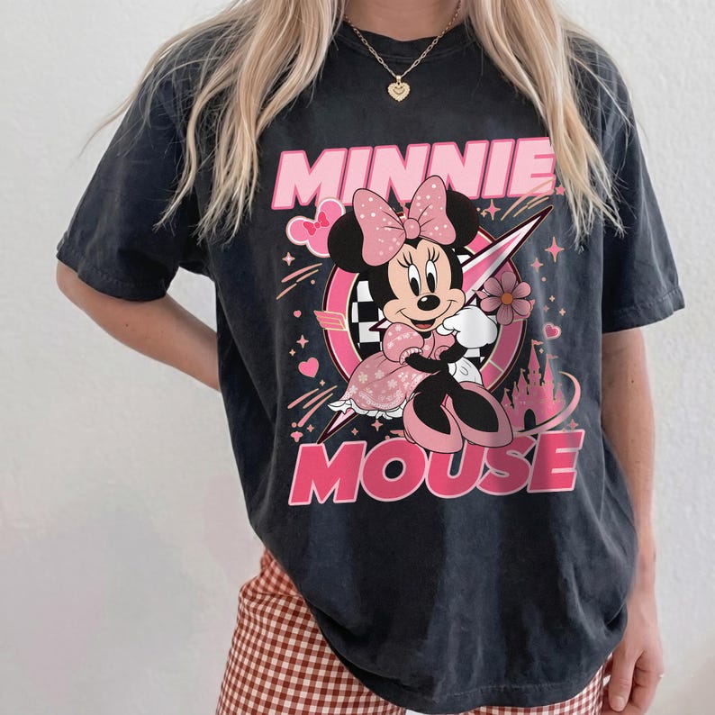 May include: Black t-shirt featuring a graphic of Minnie Mouse in a pink dress and bow. The words "MINNIE MOUSE" are printed in pink. The design includes hearts, stars, and a castle.