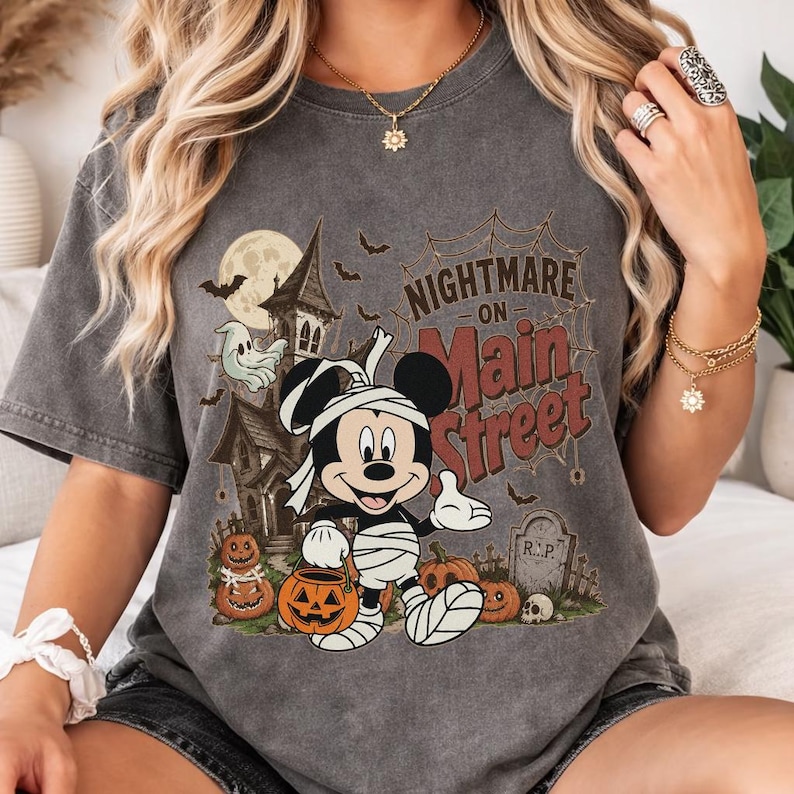 May include: Dark gray t-shirt featuring a Halloween-themed graphic. The design includes a cartoon Mickey Mouse wrapped in bandages, holding a jack-o'-lantern, with the text "Nightmare on Main Street" above a spooky scene with a haunted house and pumpkins.