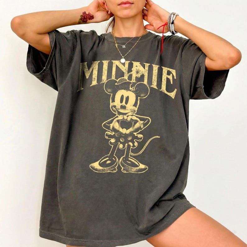 May include: Dark gray oversized t-shirt featuring a distressed graphic of Minnie Mouse with the word "MINNIE" in gold lettering. The vintage-style tee has short sleeves and a relaxed fit, perfect for casual wear.