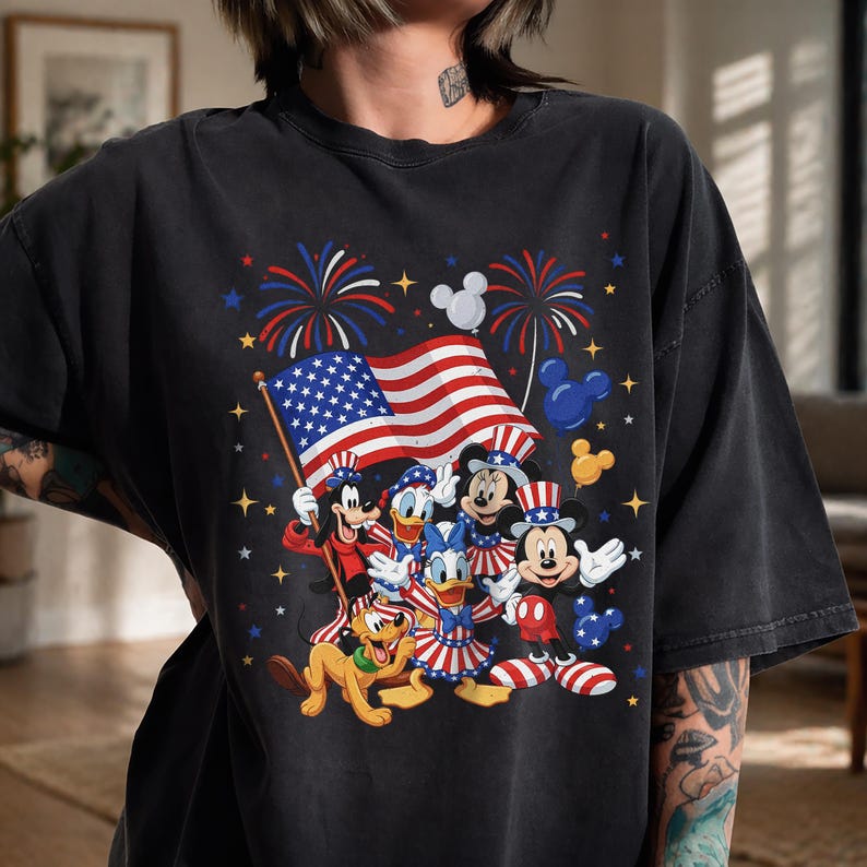 May include: Black t-shirt featuring a patriotic design with Disney characters. The graphic includes Mickey Mouse, Minnie Mouse, Donald Duck, and Pluto, along with the American flag, fireworks, and stars.