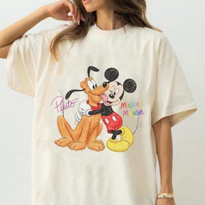 May include: Cream-coloured t-shirt with a colourful illustration of Mickey Mouse hugging Pluto. Mickey Mouse is wearing red shorts, yellow shoes, and white gloves. Pluto is light brown. The words "Pluto" and "Mickey Mouse" are written on the shirt.