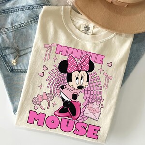 May include: Cream-coloured t-shirt featuring a graphic of Minnie Mouse in a pink dress and bow, with the words "MINNIE MOUSE" in pink. The shirt is paired with blue jeans and a tan hat.