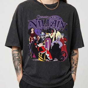 K&ouml;nnte beinhalten: Ein anthrazitfarbenes Kurzarm-T-Shirt mit einem Grafikdruck verschiedener animierter B&ouml;sewichte. Der Text "CHILLIN LIKE A VILLAIN" ist in Lila &uuml;ber den Charakteren aufgedruckt, die Outfits in Rot, Schwarz, Lila und Gelb tragen.