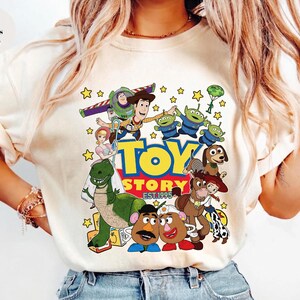 Pu&ograve; includere: T-shirt color avorio con una grafica colorata dei personaggi del film d'animazione "Toy Story". Il design include Woody, Buzz Lightyear e altri personaggi, con le parole "Toy Story EST. 1995."