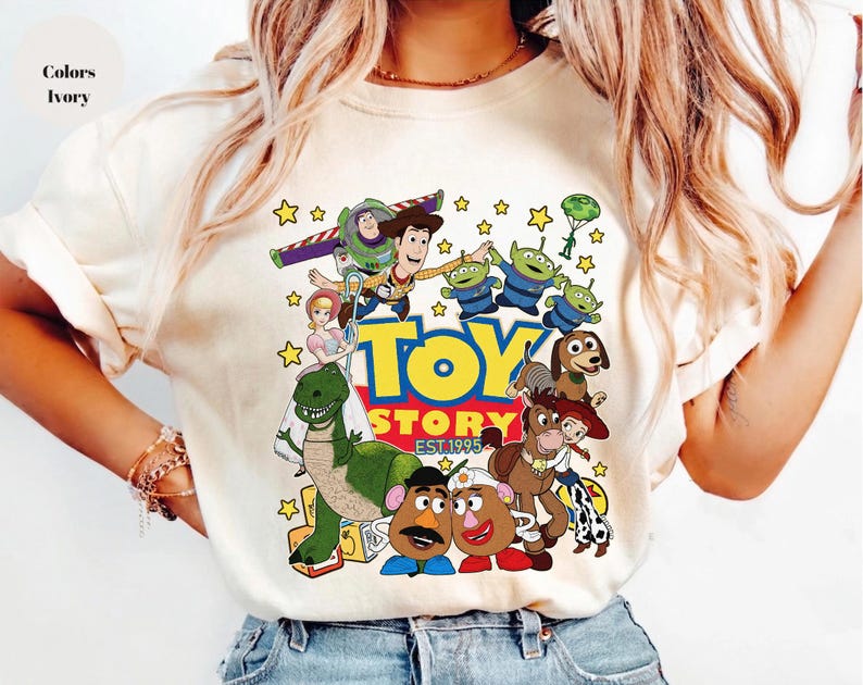 Pu&ograve; includere: T-shirt color avorio con una grafica colorata dei personaggi del film d'animazione "Toy Story". Il design include Woody, Buzz Lightyear e altri personaggi, con le parole "Toy Story EST. 1995."