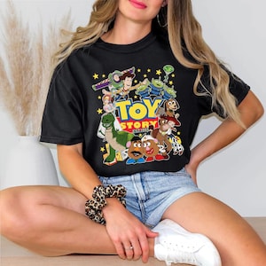 Pu&ograve; includere: T-shirt nera con una grafica colorata dei personaggi del film "Toy Story". Il design include Woody, Buzz Lightyear, gli alieni e il testo "TOY STORY EST. 1995". La maglietta &egrave; indossata da una persona.