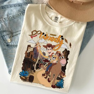 May include: Cream-colored t-shirt featuring a graphic of Sheriff Woody from Toy Story riding a horse. The design includes the words "Woody Sheriff" in a western-style font, with a town backdrop and blue accents.
