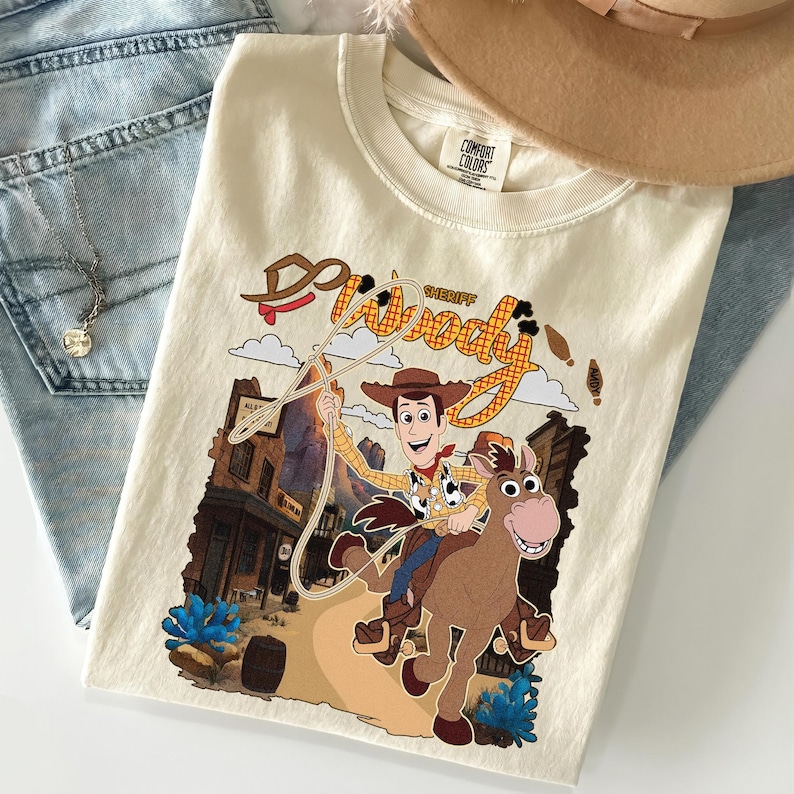 May include: Cream-colored t-shirt featuring a graphic of Sheriff Woody from Toy Story riding a horse. The design includes the words "Woody Sheriff" in a western-style font, with a town backdrop and blue accents.