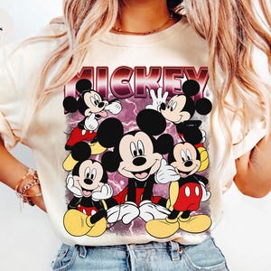 Puede incluir: Camiseta de color marfil con un gr&aacute;fico de Mickey Mouse en varias poses. La palabra "MICKEY" se muestra en una fuente retro burdeos sobre los personajes de dibujos animados. La camiseta tiene mangas cortas.