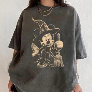 Puede incluir: Camiseta gris oscuro con una ilustraci&oacute;n beige de un personaje de dibujos animados con tem&aacute;tica de brujas sosteniendo una escoba. El personaje lleva un sombrero de bruja y un vestido. La camiseta tiene mangas cortas y un corte holgado.