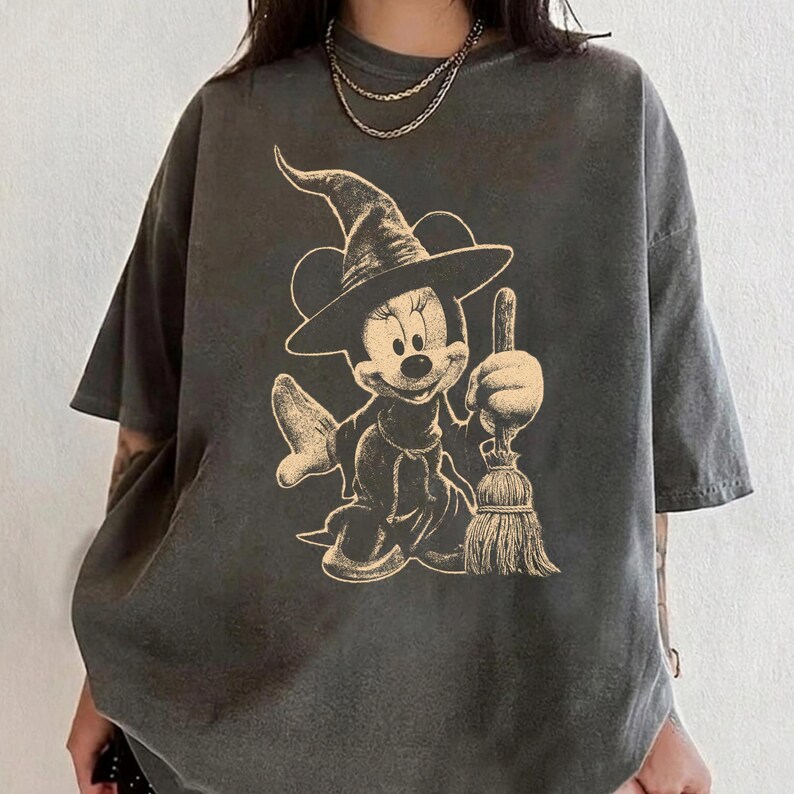 Puede incluir: Camiseta gris oscuro con una ilustraci&oacute;n beige de un personaje de dibujos animados con tem&aacute;tica de brujas sosteniendo una escoba. El personaje lleva un sombrero de bruja y un vestido. La camiseta tiene mangas cortas y un corte holgado.