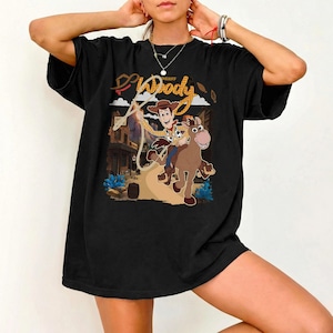 May include: Black t-shirt featuring a cartoon graphic of Sheriff Woody riding a horse with the text "Woody Sheriff". The design includes a rope, a town backdrop, and blue accents. A fun, casual tee.