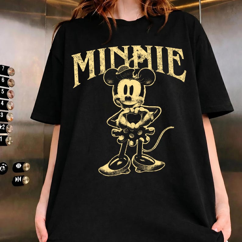 May include: Black t-shirt featuring a distressed gold graphic of Minnie Mouse. The word "MINNIE" is arched above the character. Minnie Mouse is depicted with her hands forming a heart shape. The shirt has short sleeves.