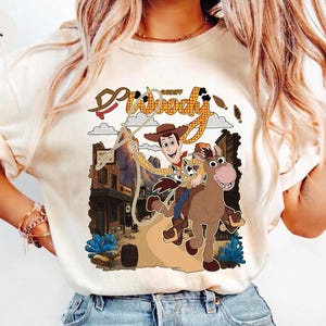 May include: Ivory-colored t-shirt featuring a graphic of Sheriff Woody from Toy Story riding a horse. The design includes the words "Sheriff Woody" in a western-style font, with a cartoon illustration of a town in the background.