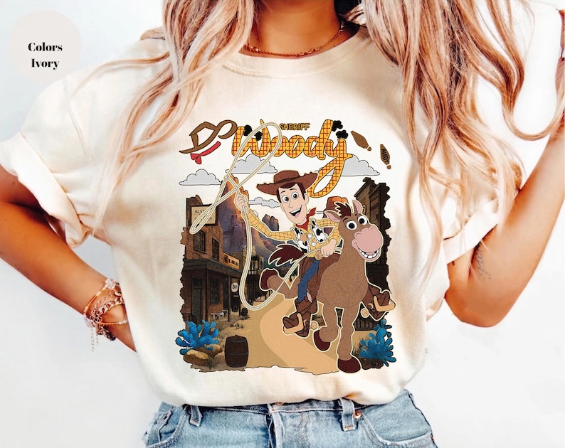 May include: Ivory-colored t-shirt featuring a graphic of Sheriff Woody from Toy Story riding a horse. The design includes the words "Sheriff Woody" in a western-style font, with a cartoon illustration of a town in the background.