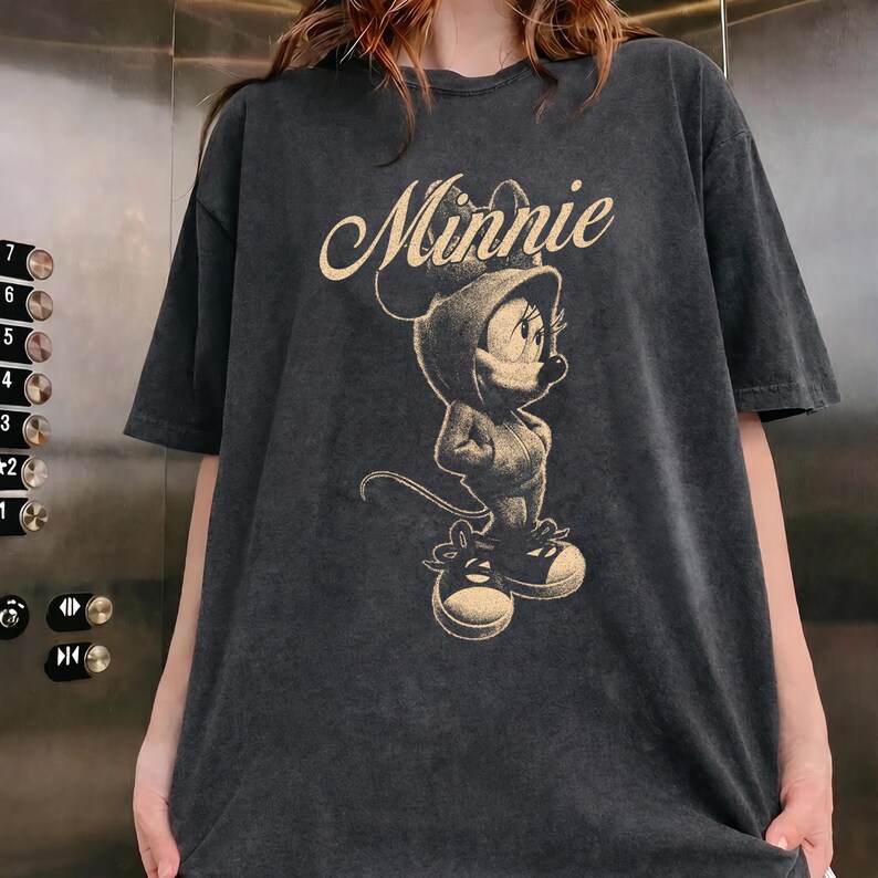 May include: A charcoal gray short-sleeve t-shirt featuring a light beige graphic print of Minnie Mouse wearing a hooded sweatshirt and sneakers. The word Minnie is printed in a cursive script above the character illustration on the front.
